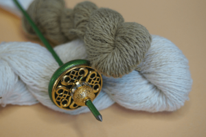 Ein Strang weiße und ein Strang grün-braune handgesponnene Baumwolle liegen übereinander vor lachsfarbenem Hintergrund. obenauf liegt eine reich verzierte Tahkli-Spindel in grün und gold.