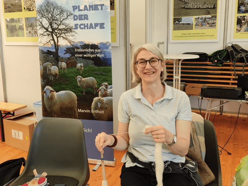 Kathrin sitzt mit Spindel und Fasern in der Hand vor einem Aufsteller "Planet der Schafe".