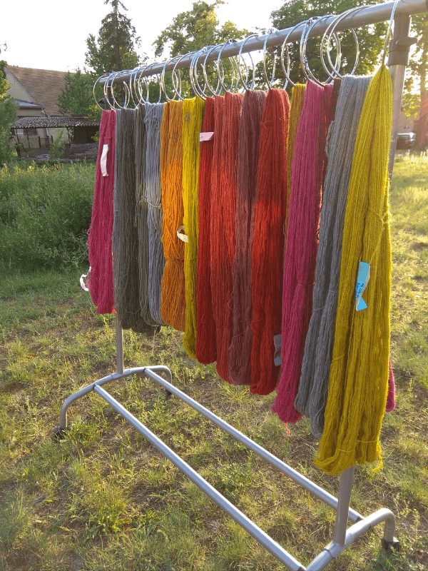 mit Naturfarben gefärbte Garne in Rot-, Gelb- und Blautönen hängen auf einer Kleiderstange zum Trocknen im sommerlichen Abendlicht.