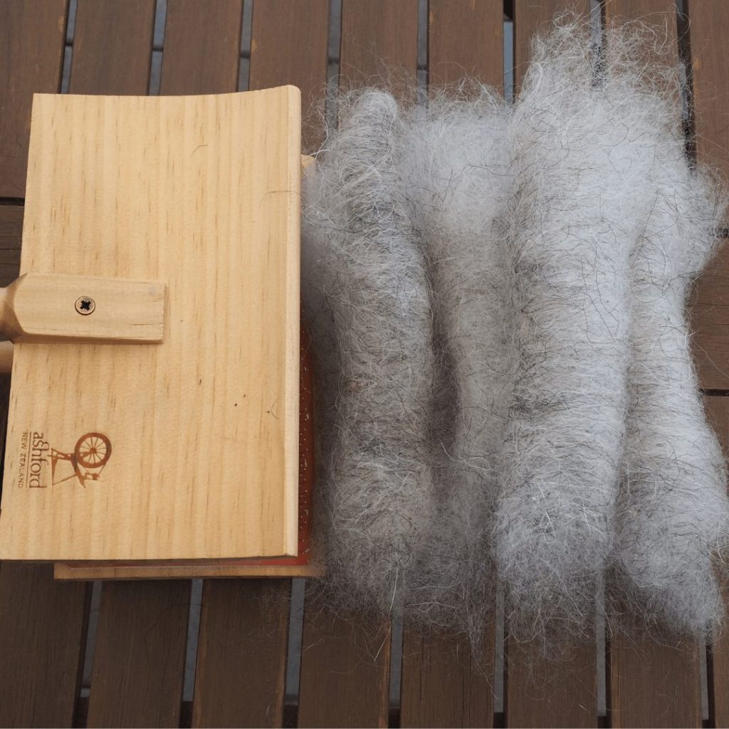 Handkardierte Rolags aus grauer Schafwolle liegen neben einer Handkarde auf einem braunen Holztisch. Aufsicht. faserexperimente.