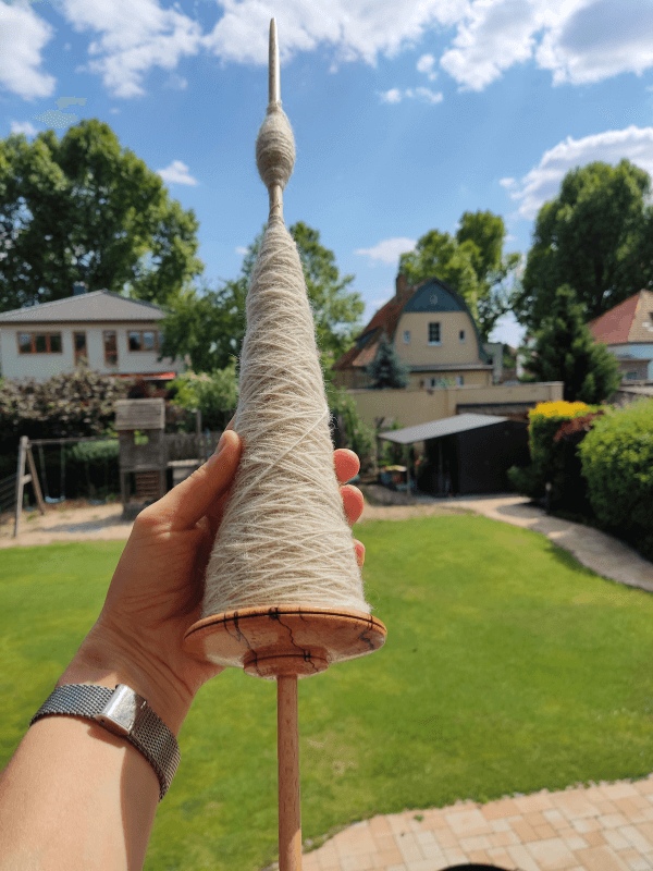 Eine Hand hält eine große Bodenspindel vor einem Hintergrund aus grüner Wiese, Spielplatz, Häusern und blauem Himmel. Die Spindel ist voll mit gesponnenem Faden.