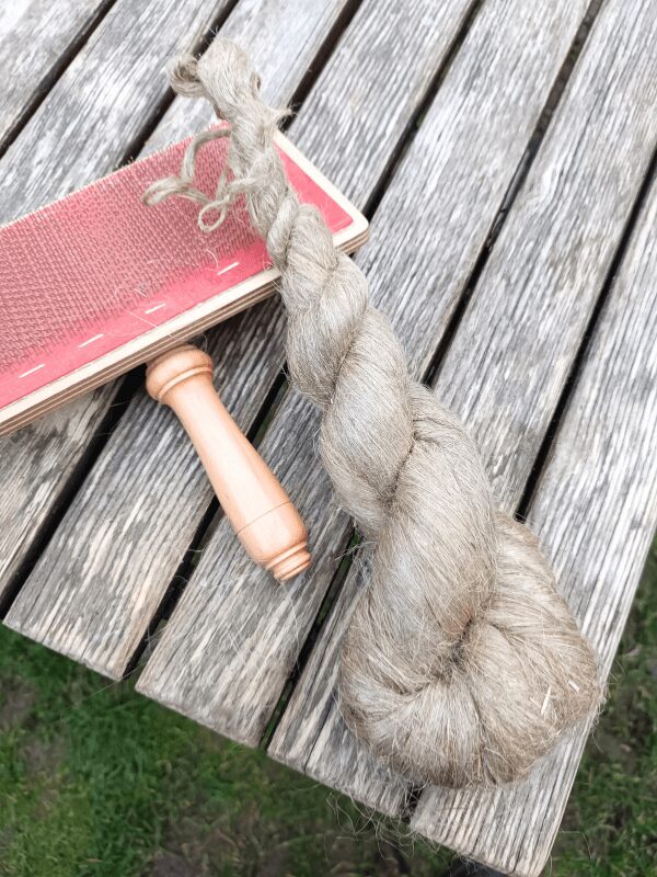 Ein Flachszopf und eine Handkarde liegen auf einem Holzlattentisch und warten auf die Verarbeitung.
