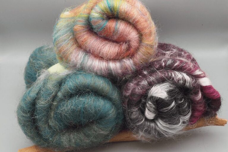 Stapel aus drei handkardierten Batts aus bunten Fasern. Die Batts sind unterschiedlich homogen, rechts unen liegt ein schwarz, weiß und rot gehaltenes Art-Batt, links unten ein sehr homogenes grünes Batt und oben ein in Pastelltönen gehaltenes Batt mit weniger Homogenistät.