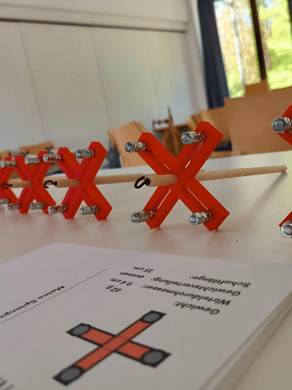 Nahaufnahme Handspindeln mit rotem 3D-gedrucktem kreuzförmigem Wirtel auf einem Tisch. Im Vordergrund liegen Karteikarten, im Hintergrund ist ein Kursraum zu sehen.