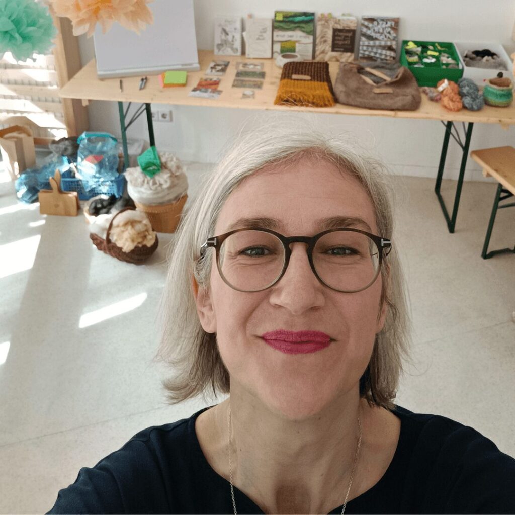 Kathrins Gesicht im Vordergrund in einem hellen Kursraum. im Hintergrund sind wollige Anschauungsmaterialien für Kursteilnehmerinnen aufgebaut.