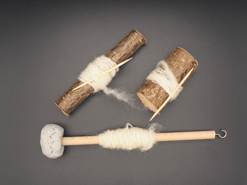 rudimentäre Spindeln bestehend aus Aststückchen mit Zahnstochern und einem Stab mit Tonklumpen als Wirtel liegen auf einem schwarzen Hintergrund. Auf allen Spindeln wurde ein Faden gesponnen.